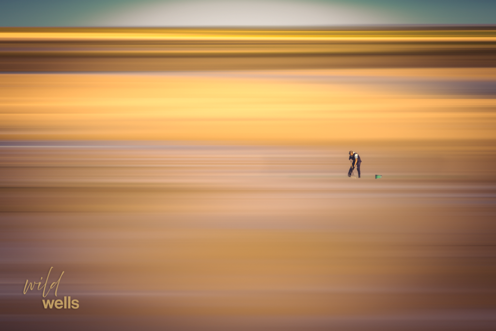 A lone figure bent forward, digging for clams on the wide tidal flats at Wells-next-the-Sea, North Norfolk, a small green bucket beside them. Photographed using the Adamski effect, the landscape dissolves into soft horizontal bands of warm amber, gold, and teal, with the figure small and right of centre against the vast, stretching sand.